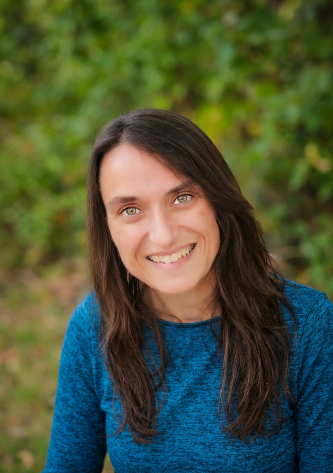 Headshot of Kristine Nemec, a smiling woman with long dark brown hair wearing a blue shirt, photographed outdoors against soft green foliage.